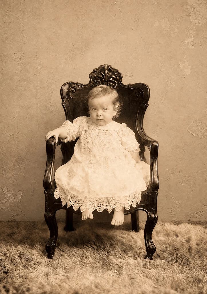 Restored studio portrait: a baby in a long lace gown seated in a carved armchair on a fur rug
