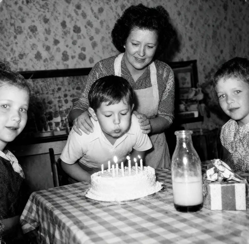 Restored birthday snapshot kept in black and white: a small boy blowing out the candles on his cake while his mother steadies him from behind