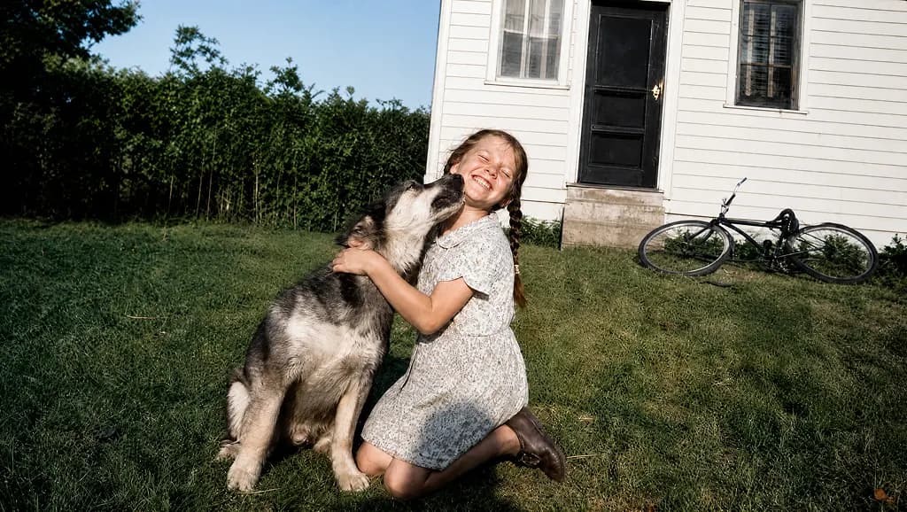 Restored snapshot: a girl in a print dress kneeling on the lawn hugging her dog, a white clapboard house and a bicycle behind them