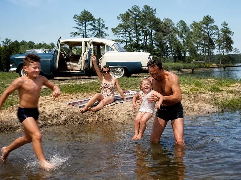 Restored summer snapshot: a father lifting his laughing little girl in the shallows of a lake while her brother runs into the water, the family car parked on the sand behind them