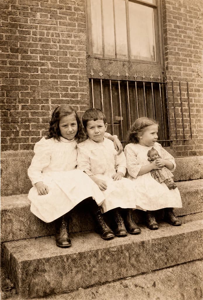 Restored photo: three young children in white dresses and leather boots sitting together on a stone stoop, the youngest holding her doll