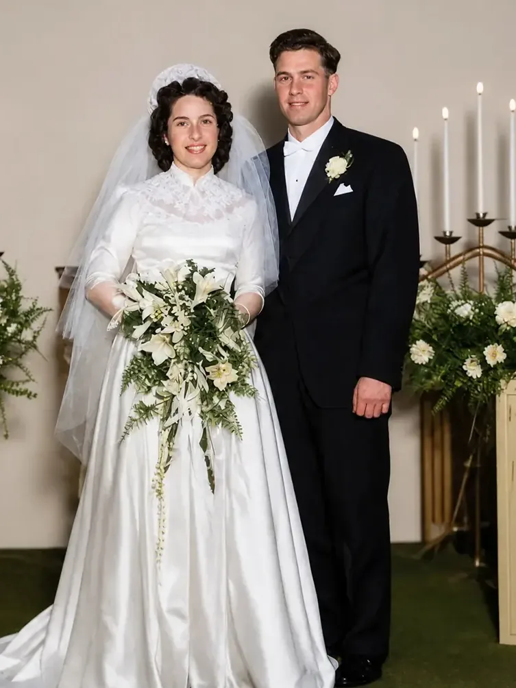 Restored wedding portrait in full color: a bride in a lace gown and veil beside her groom at a candlelit altar banked with white flowers